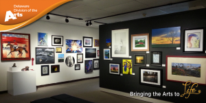 Delaware Division of the Arts orange banner over an image of a previous Delaware State Employee Art Exhibition in the Delaware State University Art Center/Gallery with multiple framed and unframed images hanging on a black wall in the foreground and white wall in the background